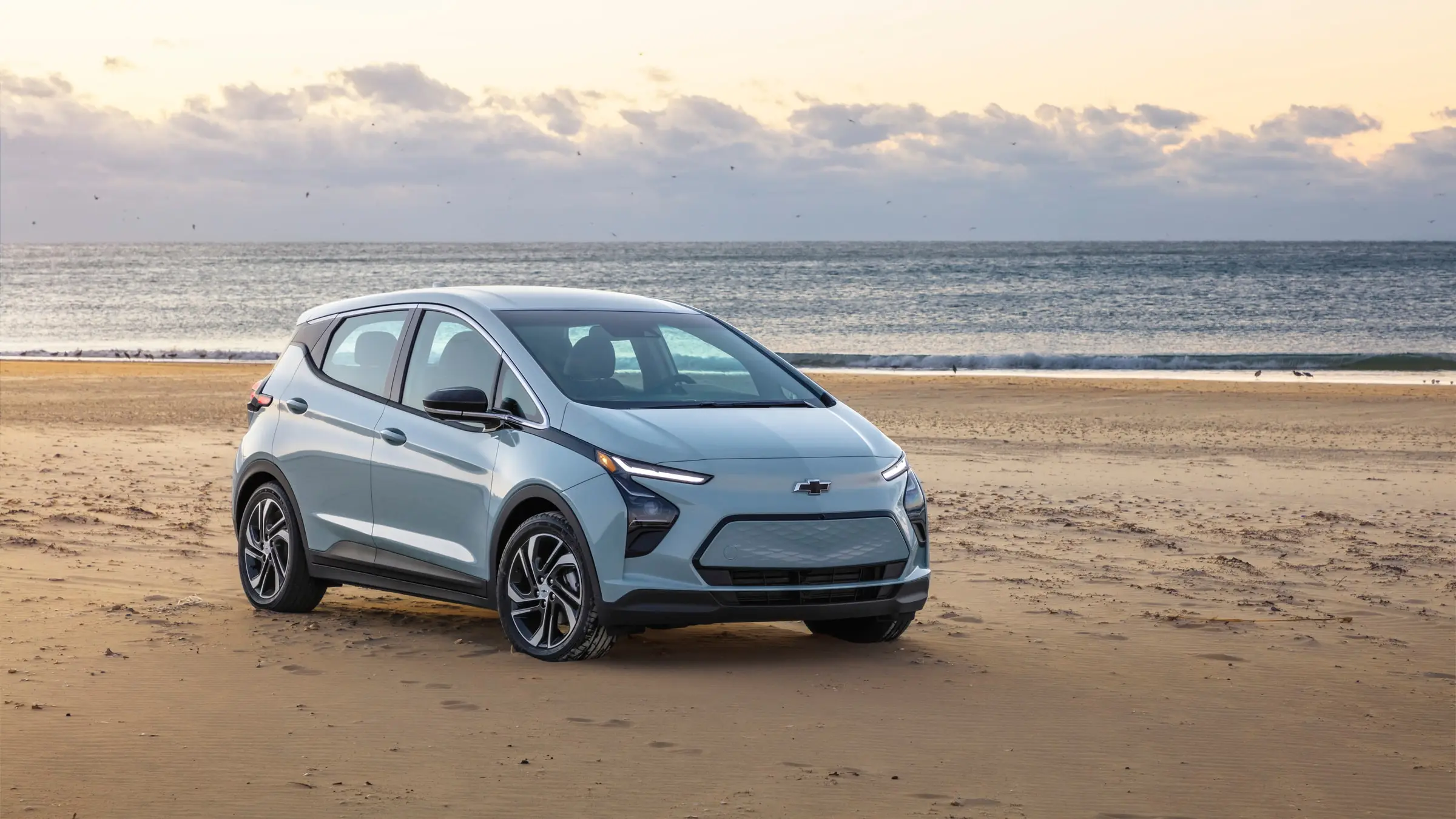 A light blue 2022 Chevrolet Bolt EV parked on a sandy beach with the ocean and a cloudy sky behind it