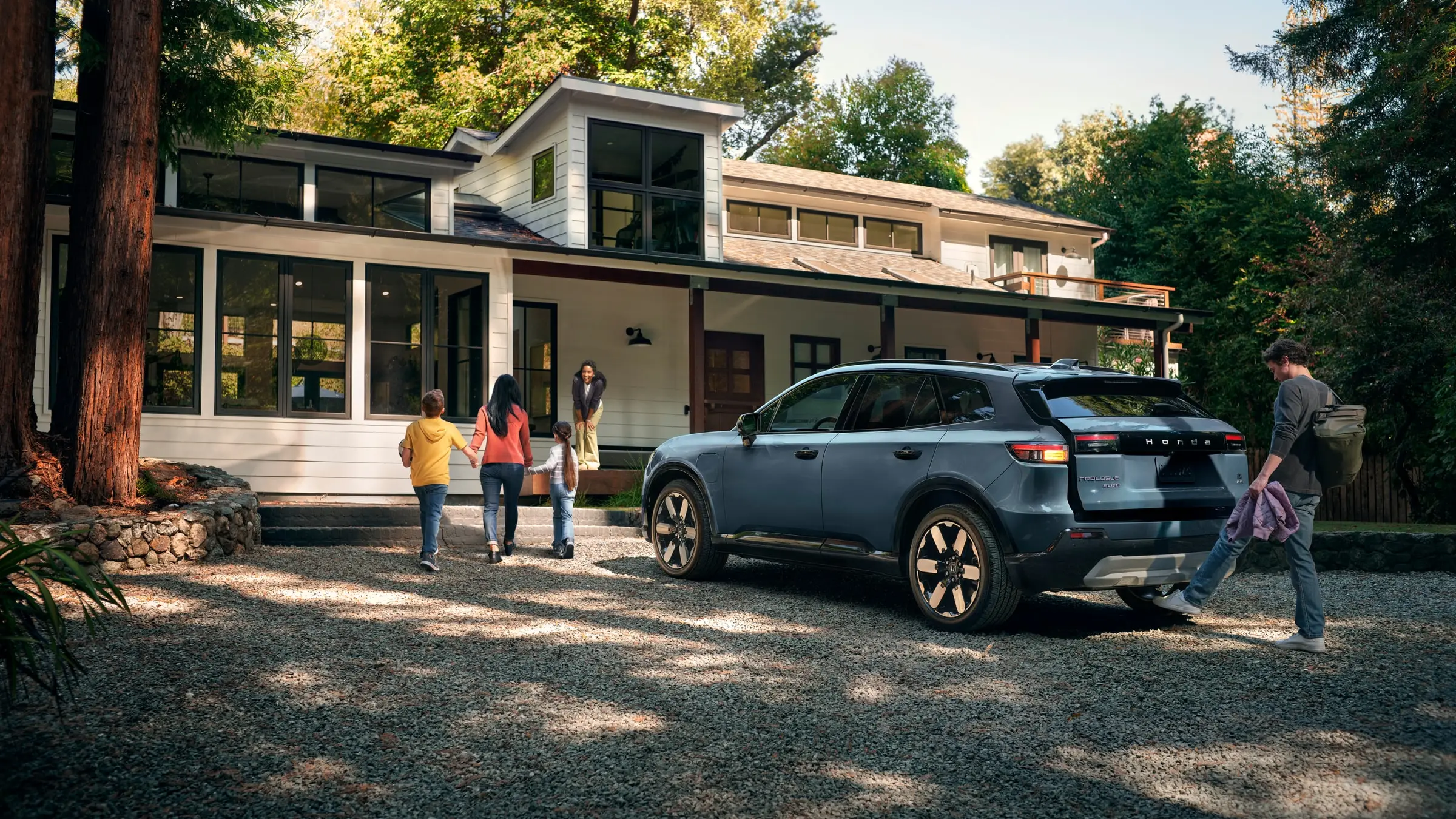A blue-grey Honda Prologue Elite parked on a gravel drive outside a white timber house as a family walks to the door