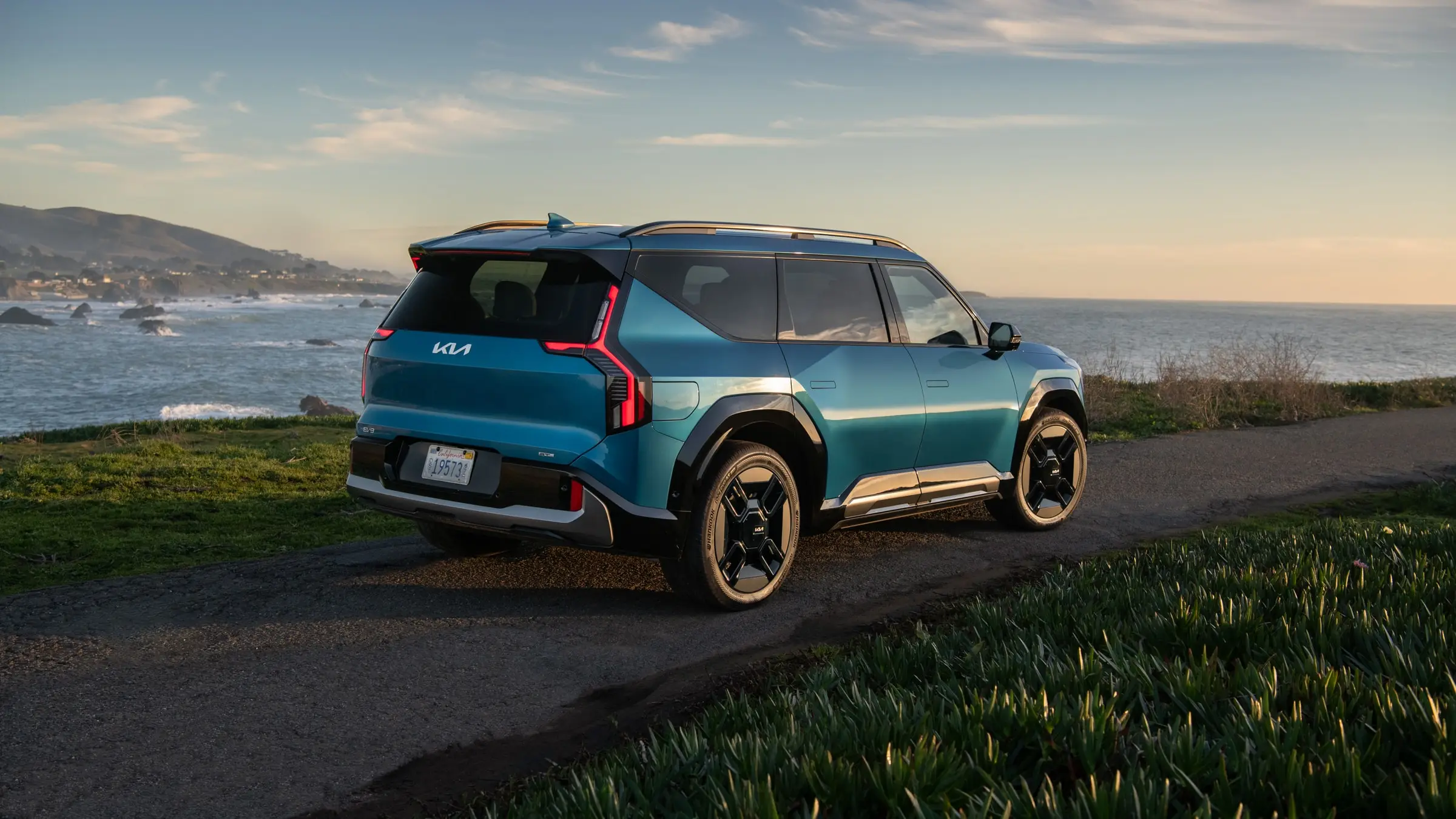 A teal-blue Kia EV9 GT-Line seen from the rear three-quarter, parked on a coastal path above a rocky shoreline at golden hour