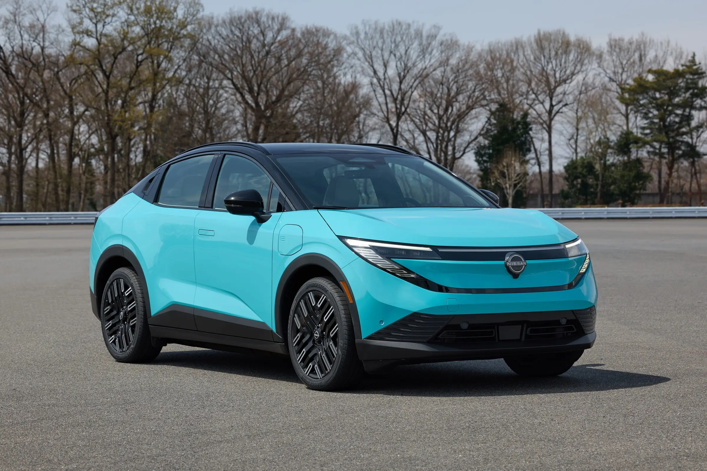 A teal 2026 Nissan Leaf parked on a test track with bare trees behind it
