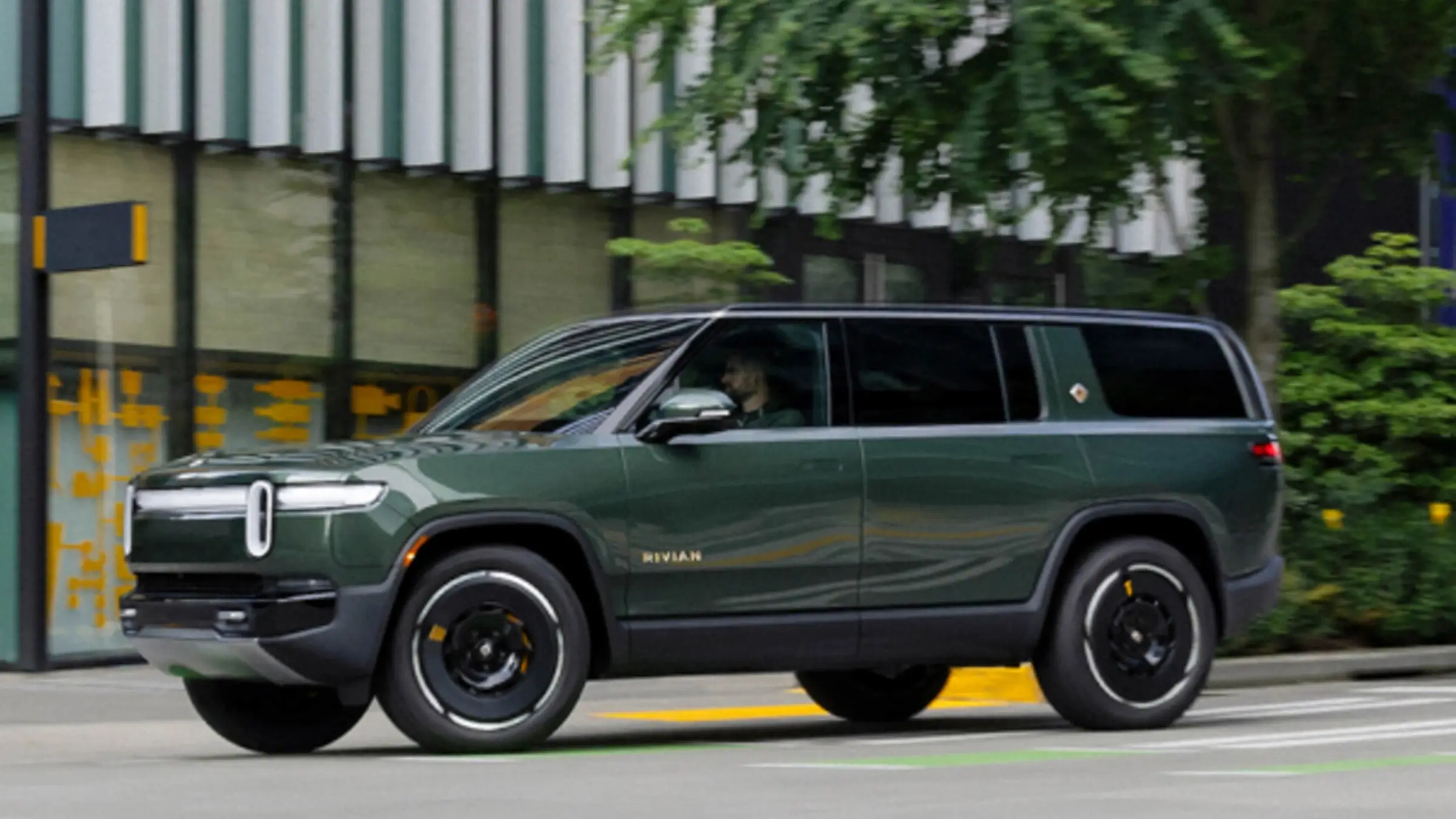 A green Rivian R1S driving past a city storefront with trees behind it