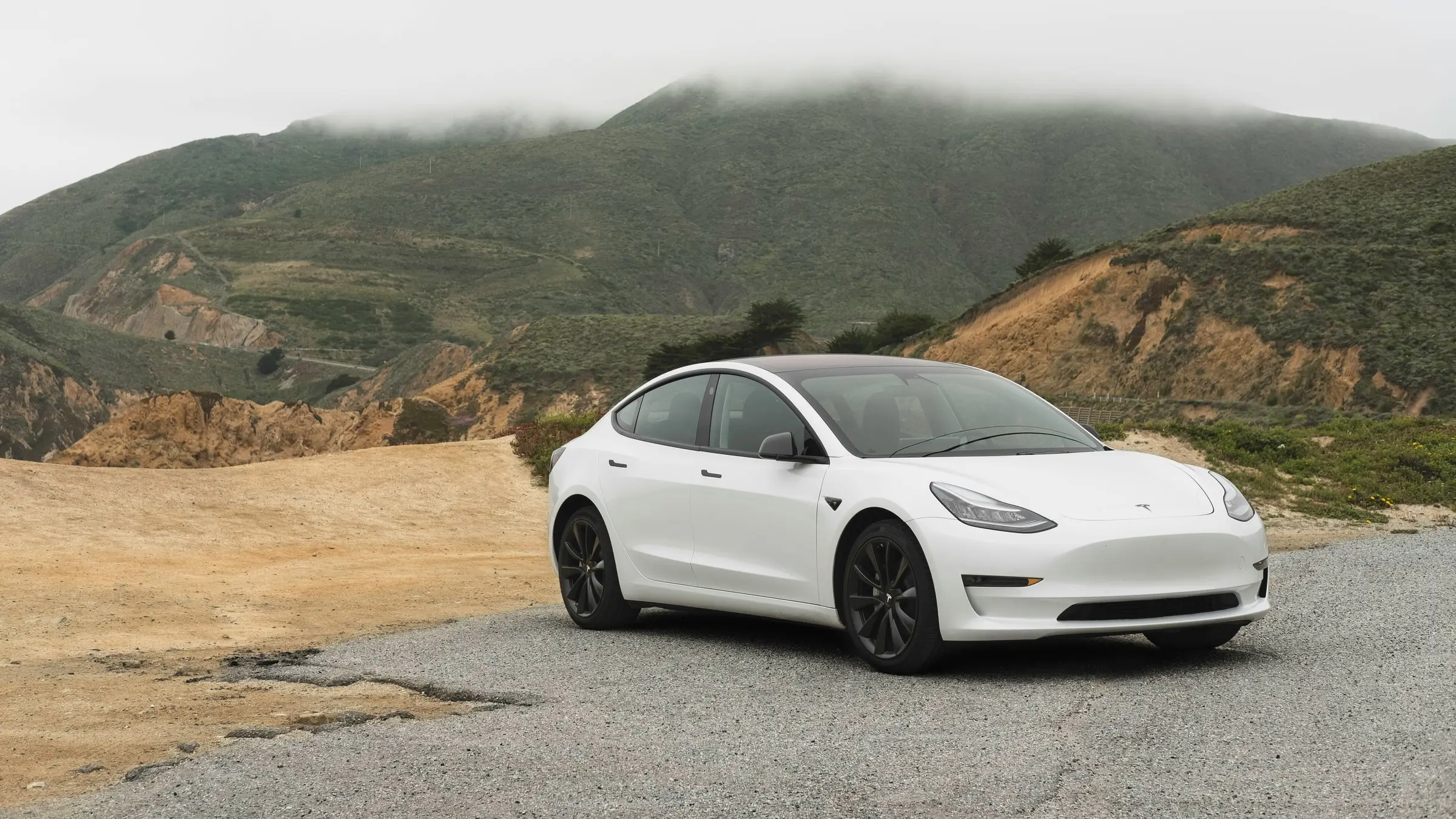 A white Tesla Model 3 parked on a gravel pull-off below a fog-topped coastal bluff
